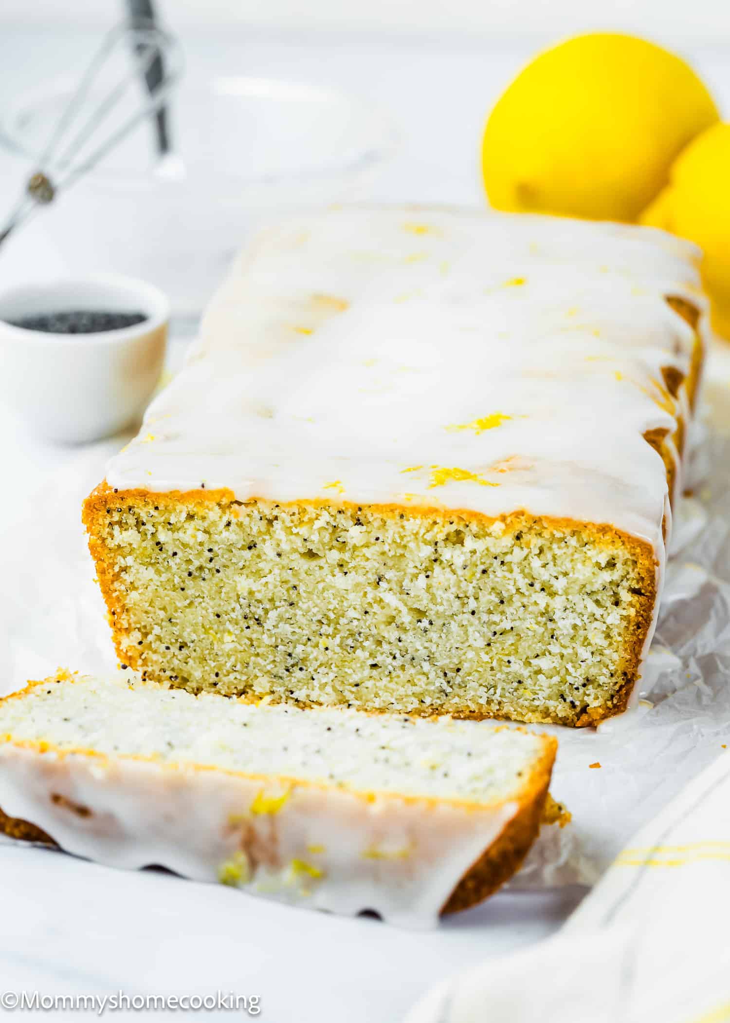 An Eggless Lemon Poppy Seed Loaf Cake with a sweet glaze, one slice cut, surrounded by fresh lemons and a small bowl of poppy seeds on a white background.