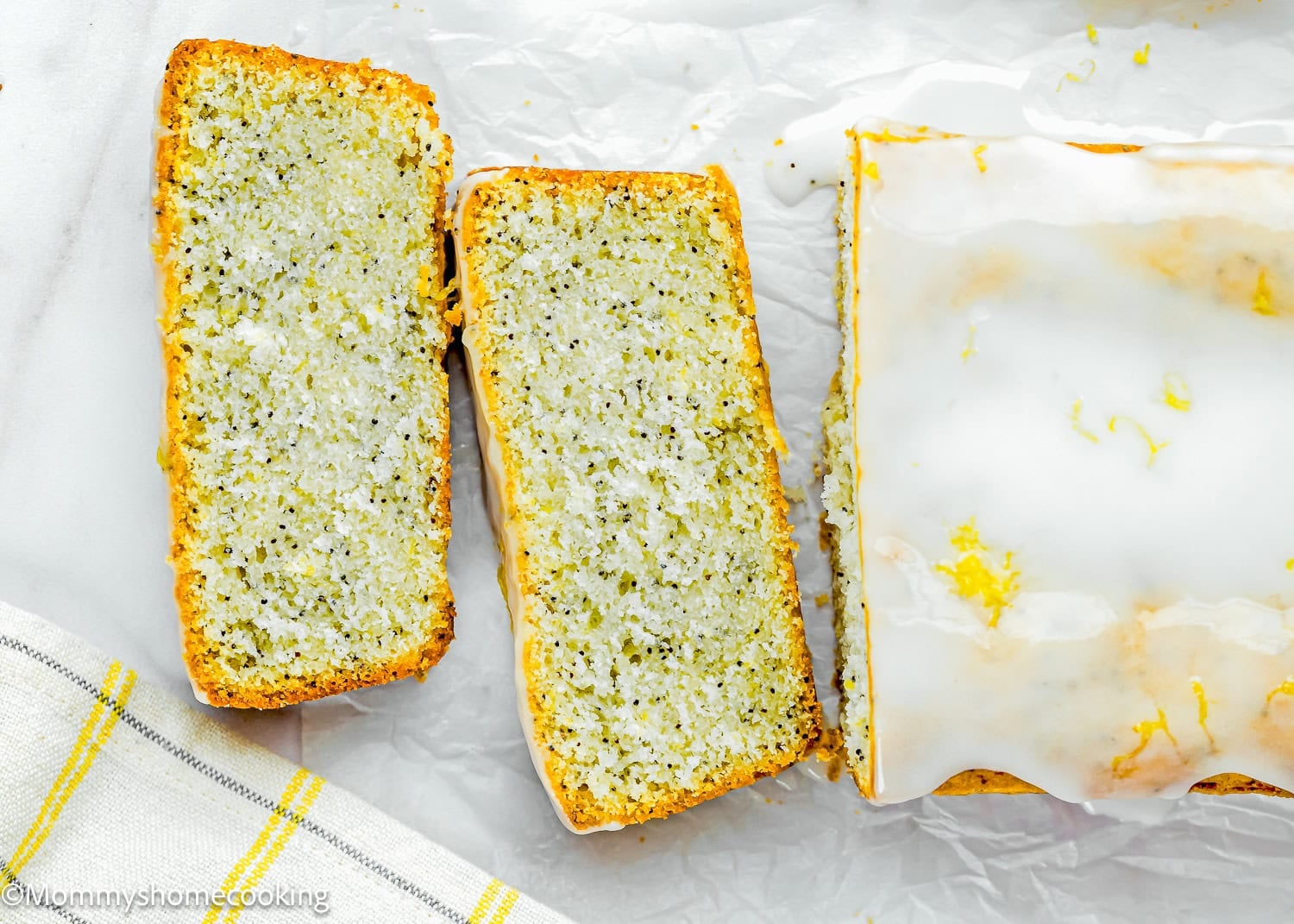 An Eggless Lemon Poppy Seed Loaf Cake with white icing, partially sliced, is displayed on parchment paper next to a striped towel.