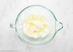 A glass mixing bowl containing white granulated sugar and lemon zest, essential for preparing an Eggless Lemon Poppy Seed Loaf Cake, sits on a white marble surface.
