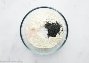 A glass bowl containing flour, salt, baking powder, and black poppy seeds for an Eggless Lemon Poppy Seed Loaf Cake sits on a white surface.
