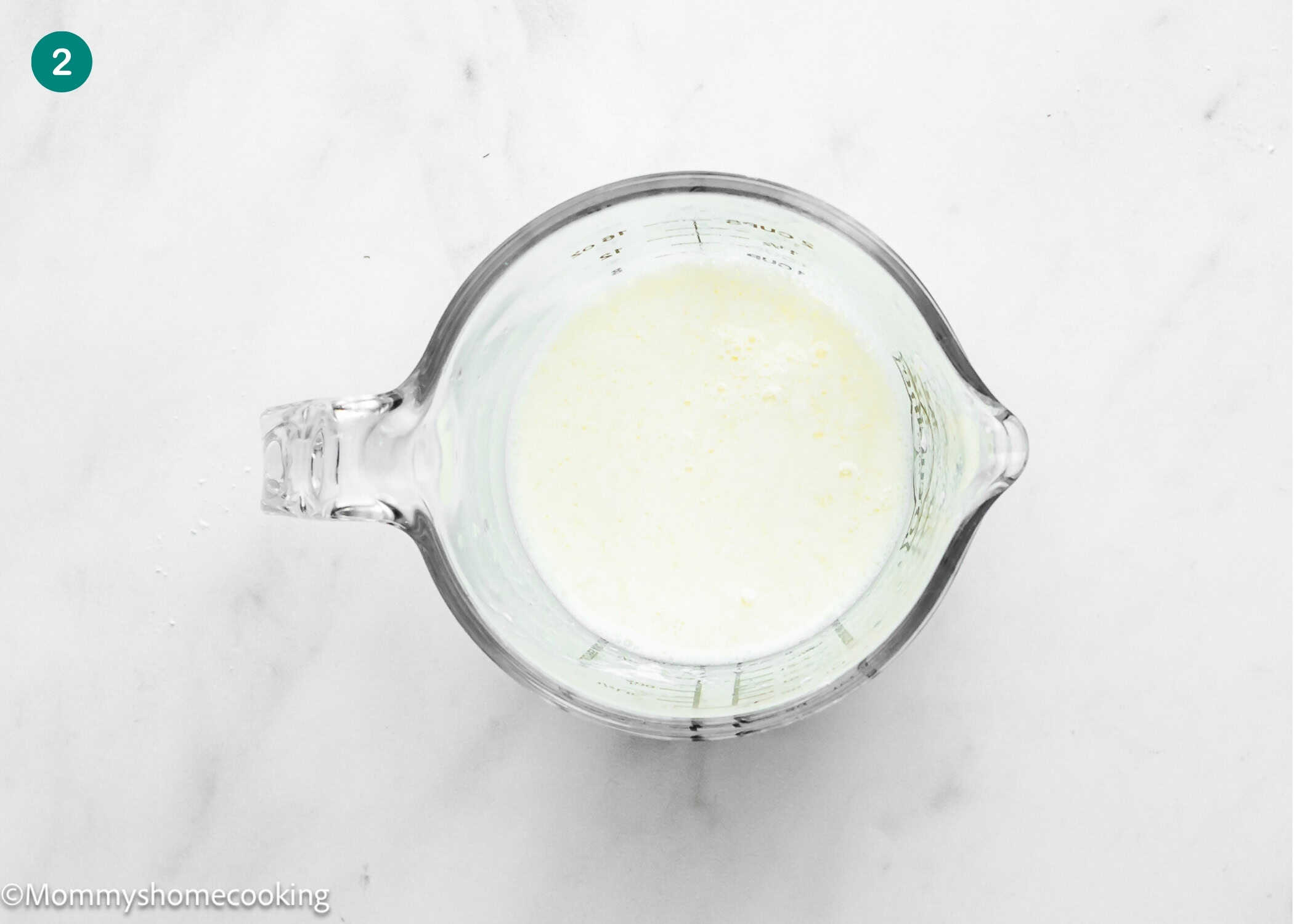 Glass measuring cup filled with a white liquid mixture for Eggless Lemon Poppy Seed Loaf Cake, placed on a white marble surface.