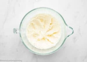 Glass bowl filled with whipped cream cheese frosting on a white marble surface, perfect for topping Eggless Lemon Poppy Seed Loaf Cake.