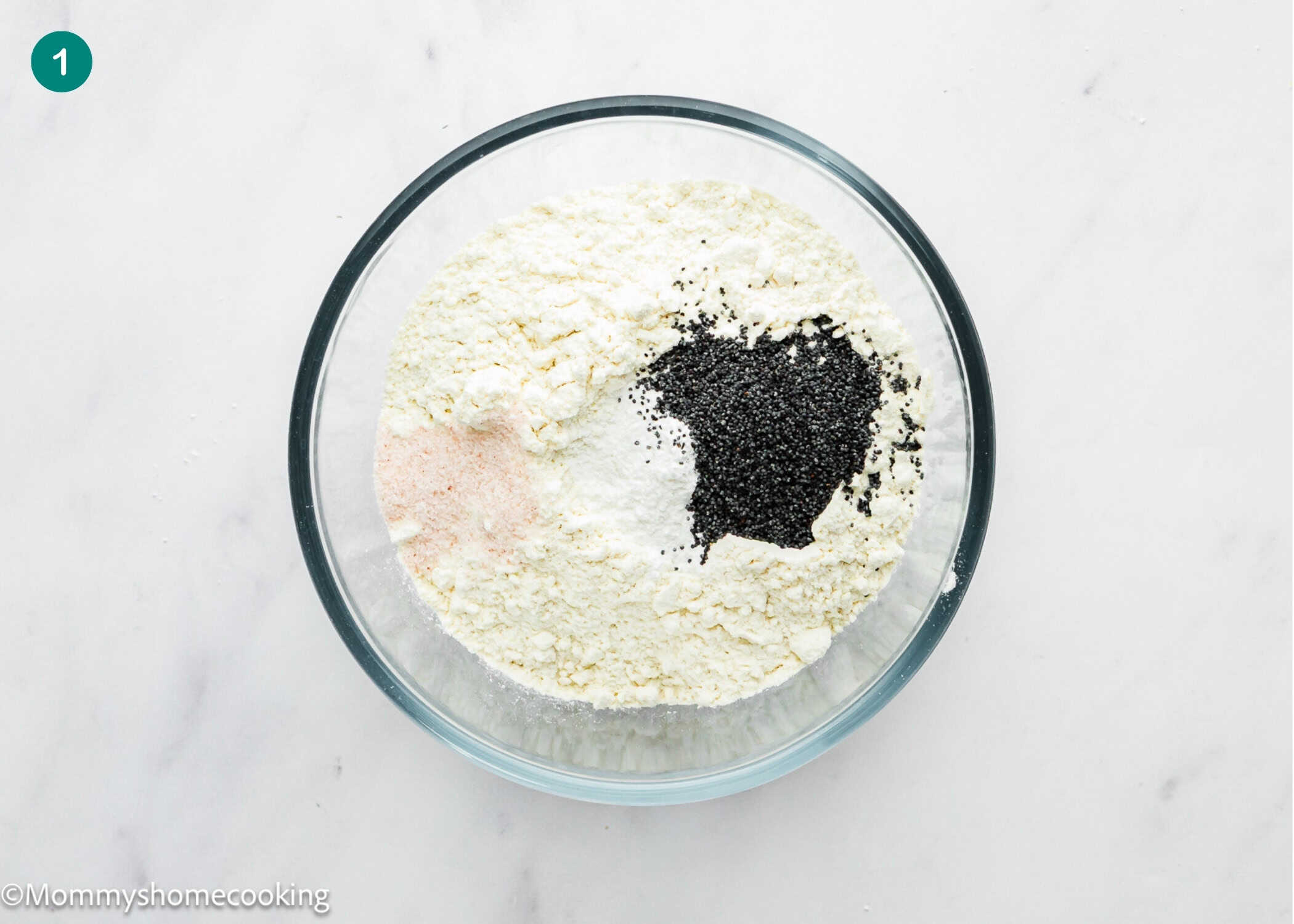 A glass bowl containing flour, baking powder, salt, and black poppy seeds for an Eggless Lemon Poppy Seed Loaf Cake on a white marble surface.