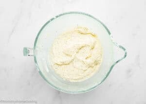 Glass mixing bowl containing a creamy mixture with visible specks, likely for an Eggless Lemon Poppy Seed Loaf Cake, on a white marble surface.