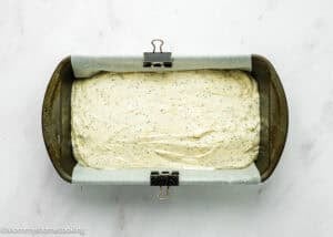 A loaf pan lined with parchment paper and filled with uncooked Eggless Lemon Poppy Seed Loaf Cake batter, secured with metal binder clips, on a white surface.