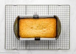 An Eggless Lemon Poppy Seed Loaf Cake rests in a parchment-lined loaf pan on a cooling rack over a white surface.
