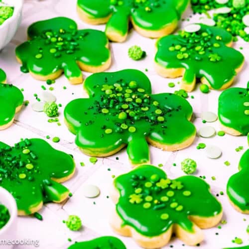 Eggless Patrick’s Day Sugar Cookies shaped like shamrocks are decorated with green icing and sprinkles, arranged on a white surface with extra festive decorations scattered around.