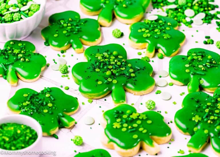 Eggless Patrick’s Day Sugar Cookies shaped like shamrocks are decorated with green icing and sprinkles, arranged on a white surface with extra festive decorations scattered around.