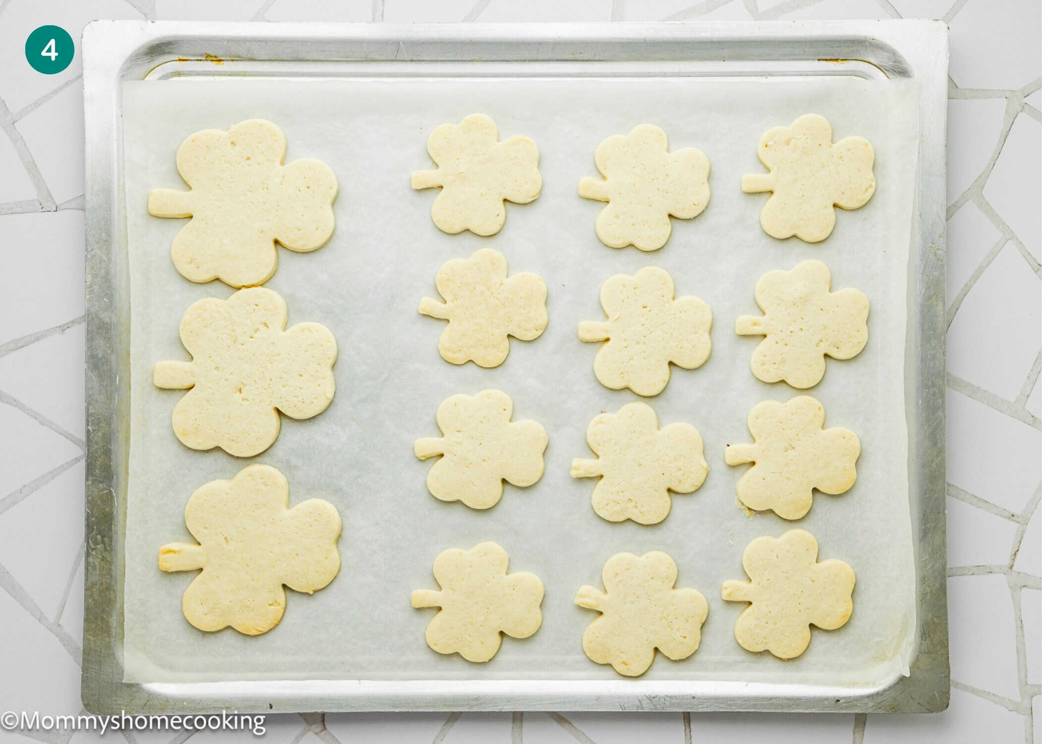 A baking sheet lined with parchment paper holds 16 unbaked shamrock-shaped Eggless Patrick’s Day Sugar Cookies ready to be baked.