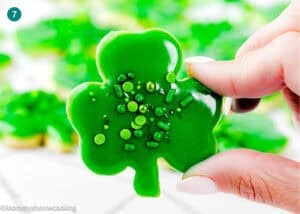 A hand holds an Eggless Patrick’s Day Sugar Cookie shaped like a shamrock with green sprinkles; more similar cookies are blurred in the background.