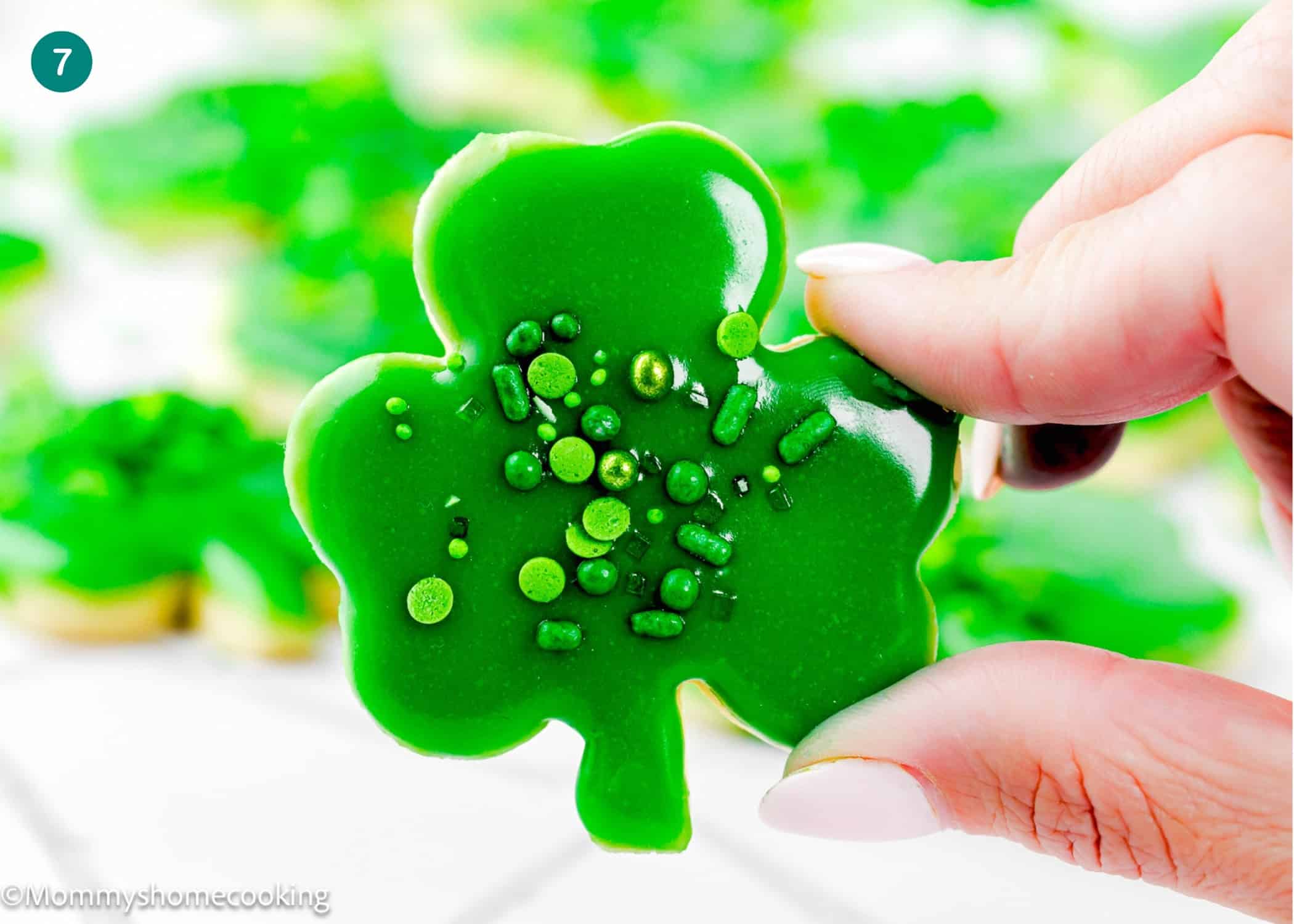 A hand holds an Eggless Patrick’s Day Sugar Cookie shaped like a shamrock with green sprinkles; more similar cookies are blurred in the background.
