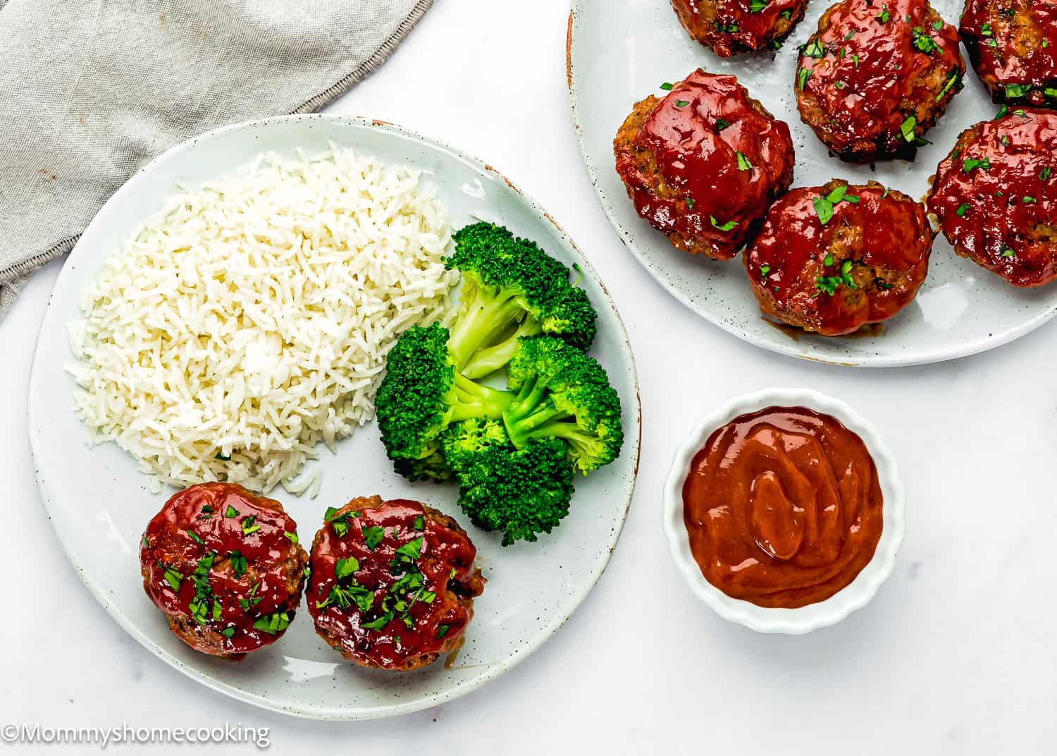 A plate with two Mini Meatloaf Muffins (No Eggs!) topped with sauce, served alongside white rice and steamed broccoli. More mini meatloaf muffins and a small bowl of sauce are nearby.