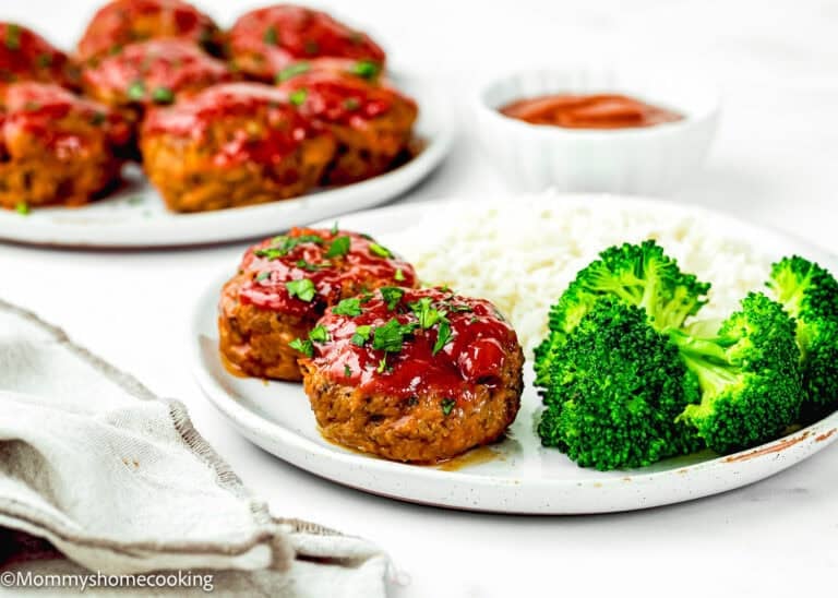 A plate with two Mini Meatloaf Muffins (No Eggs!) topped with sauce, served with steamed broccoli and white rice. Extra mini meatloaf muffins and sauce are in the background.