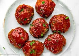 Seven glazed Mini Meatloaf Muffins (No Eggs!) are arranged on a white plate, garnished with chopped parsley.