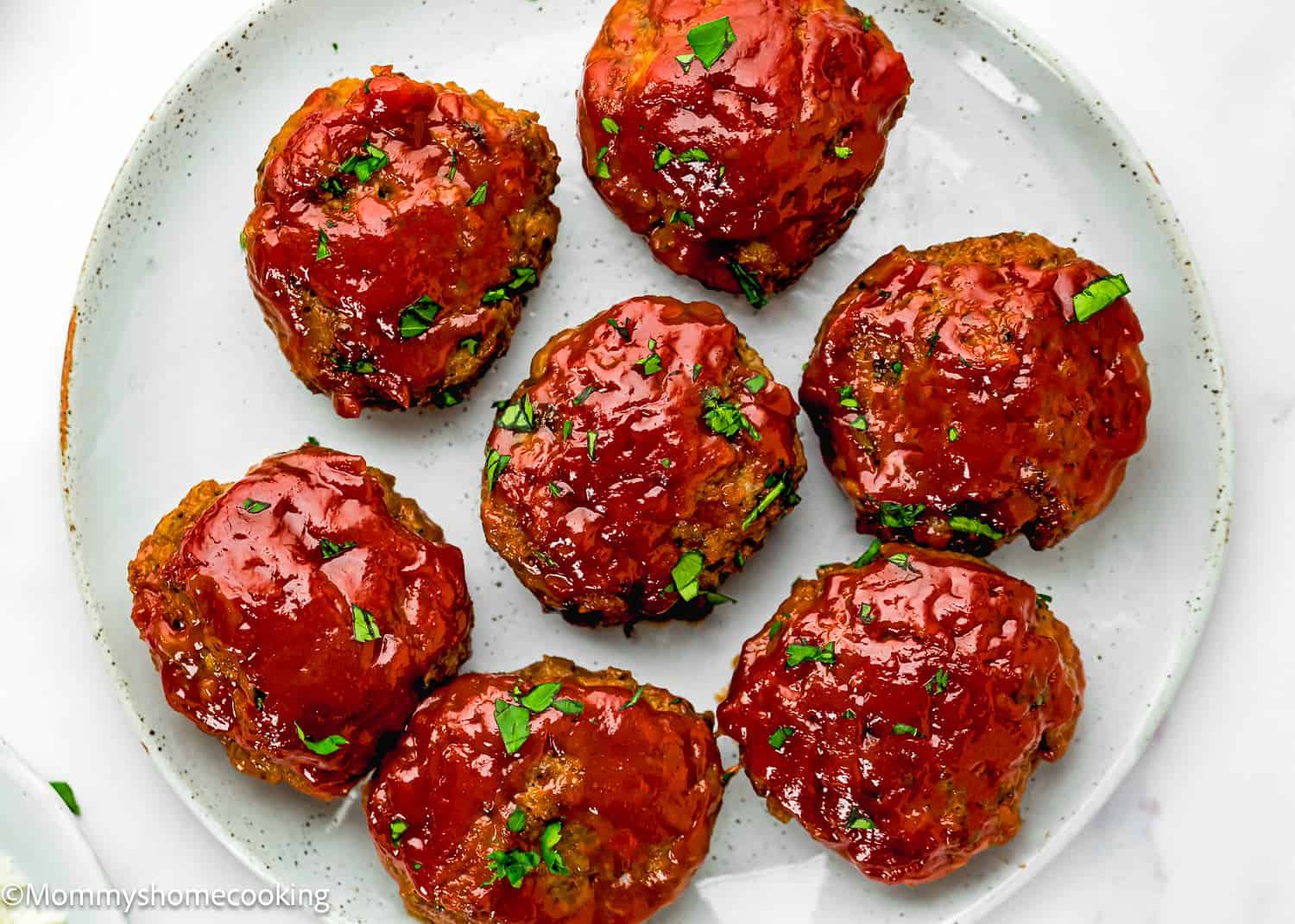 Seven glazed Mini Meatloaf Muffins (No Eggs!) are arranged on a white plate, garnished with chopped parsley.