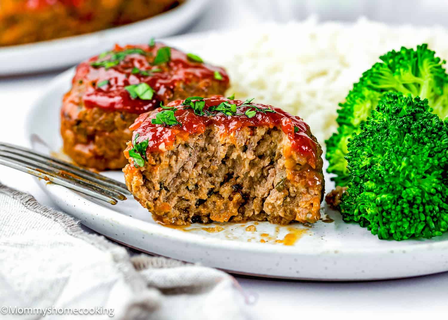 A plate with two Mini Meatloaf Muffins (No Eggs!) topped with ketchup and herbs, served with steamed broccoli and white rice. A fork rests on the plate.