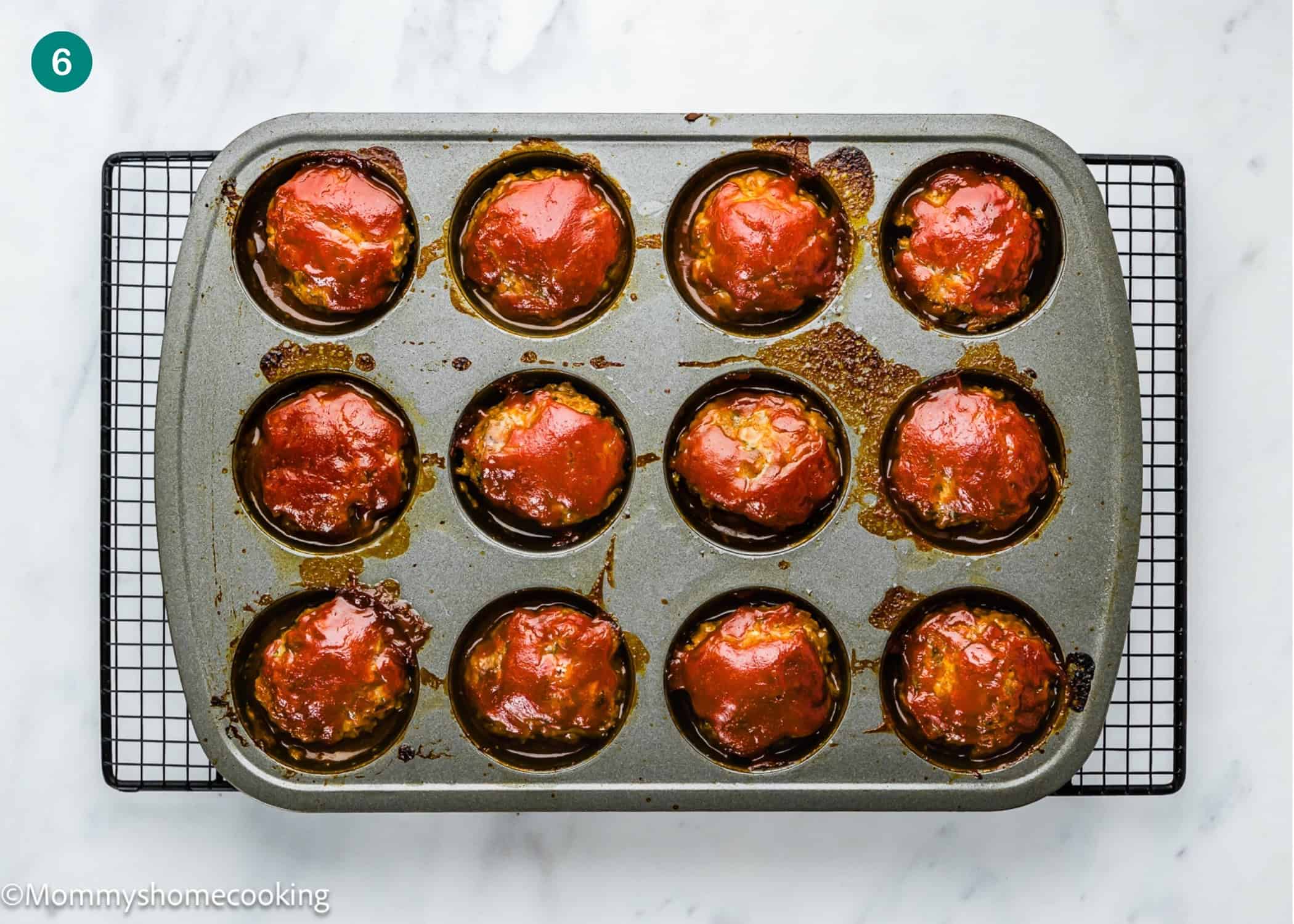 A muffin tin filled with twelve Mini Meatloaf Muffins (No Eggs!) topped with red sauce, resting on a cooling rack over a marble surface.