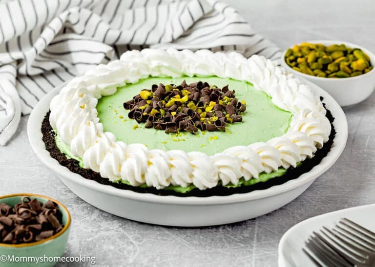 A No Bake Pistachio Chocolate Cheesecake Pie with a mint green filling, whipped cream border, chocolate curls, and chopped pistachios sits on a white plate next to bowls of chocolate and pistachios.