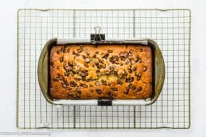 A loaf of One Bowl Egg-Free and Dairy-Free Chocolate Chip Banana Bread in a baking pan lined with parchment paper, cooling on a wire rack.