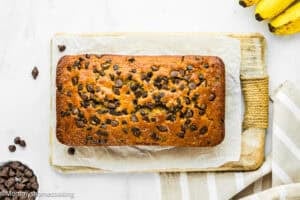 A loaf of One Bowl Egg-Free and Dairy-Free Chocolate Chip Banana Bread sits on parchment paper atop a wooden cutting board, with chocolate chips and bananas nearby.