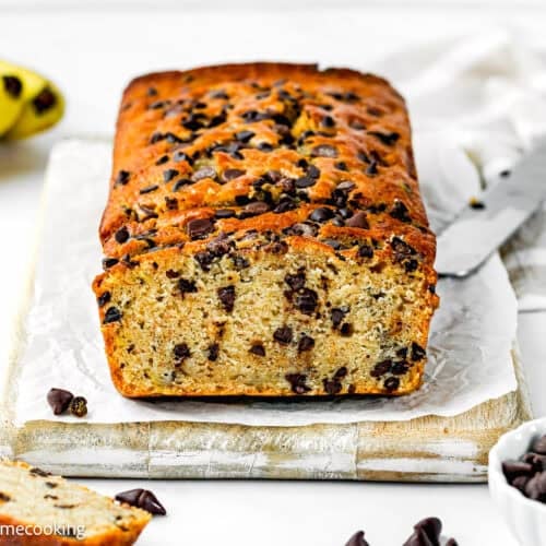 A loaf of One Bowl Egg-Free and Dairy-Free Chocolate Chip Banana Bread sits on parchment paper, with a knife and a bowl of chocolate chips nearby.