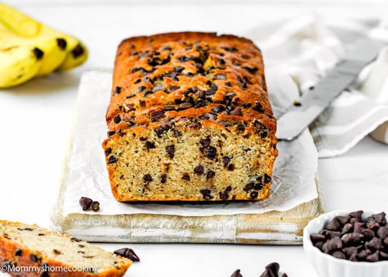 A loaf of One Bowl Egg-Free and Dairy-Free Chocolate Chip Banana Bread sits on parchment paper, with a knife and a bowl of chocolate chips nearby.