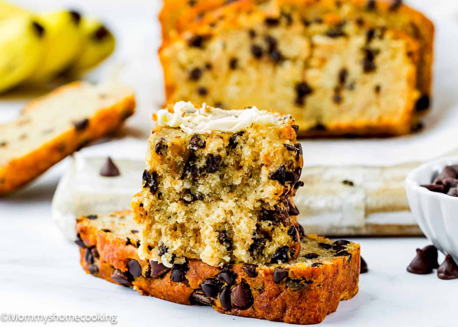 Slices of One Bowl Egg-Free and Dairy-Free Chocolate Chip Banana Bread are stacked on a table, with the top piece spread with butter. A bowl of chocolate chips and bananas are in the background.