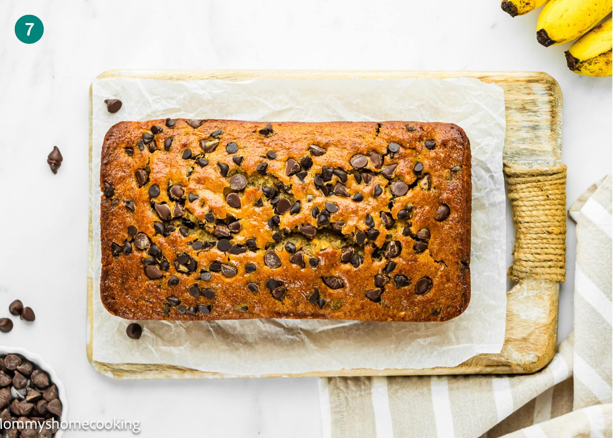 A loaf of One Bowl Egg-Free and Dairy-Free Chocolate Chip Banana Bread sits on parchment paper on a wooden cutting board, with bananas and a bowl of chocolate chips nearby.