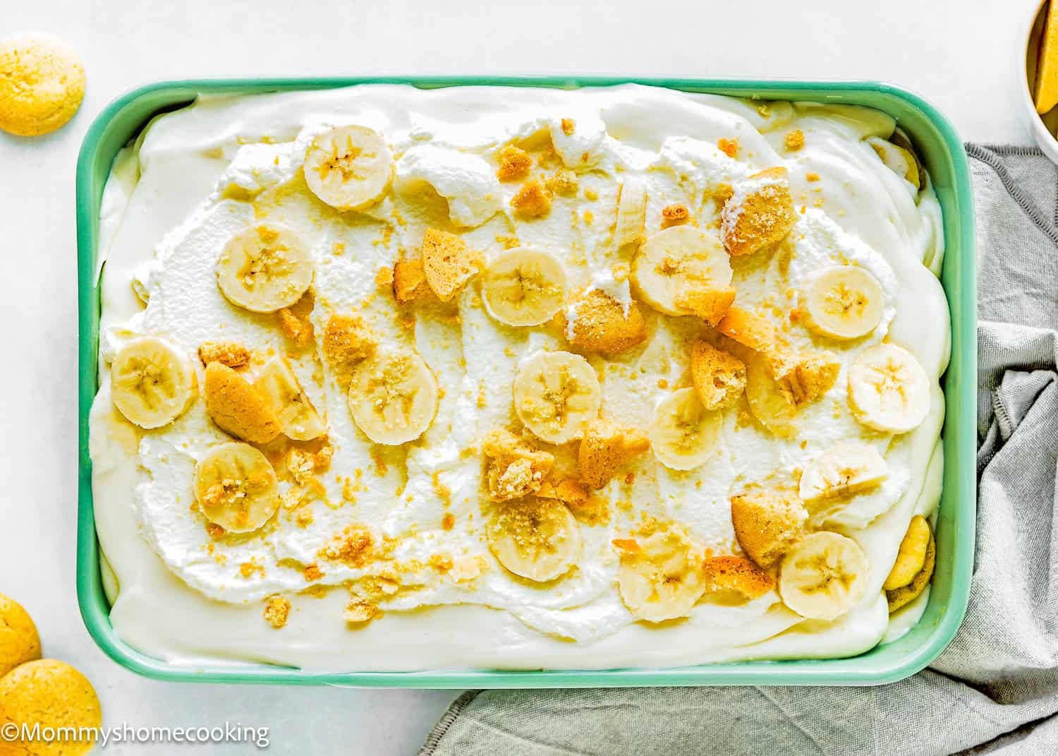 Rectangular dish filled with The Best Eggless Banana Pudding Recipe, topped with whipped cream, banana slices, and crumbled vanilla wafers. A few whole vanilla wafers are placed beside the dish.