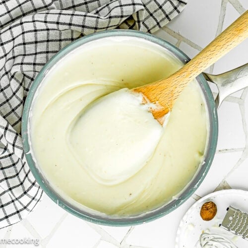 A saucepan filled with Easy Béchamel Sauce (Classic White Sauce Recipe) is being stirred with a wooden spoon, next to a checkered cloth and a small plate holding a grater and nutmeg.