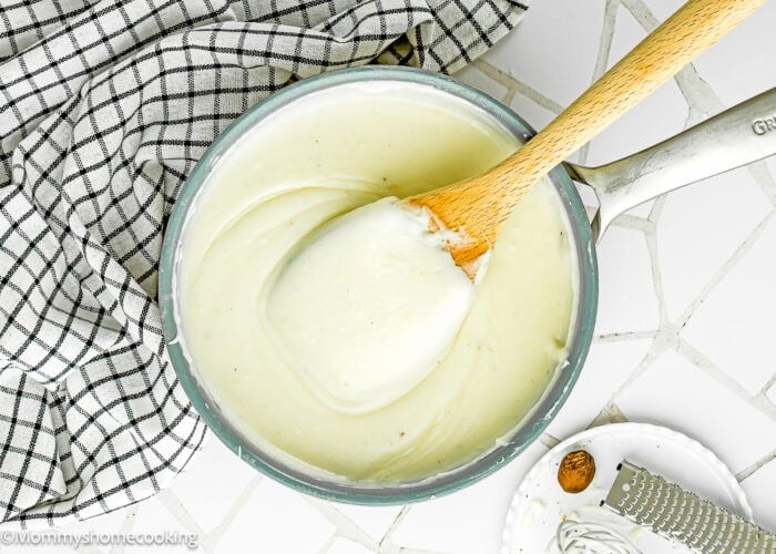 A saucepan filled with Easy Béchamel Sauce (Classic White Sauce Recipe) is being stirred with a wooden spoon, next to a checkered cloth and a small plate holding a grater and nutmeg.
