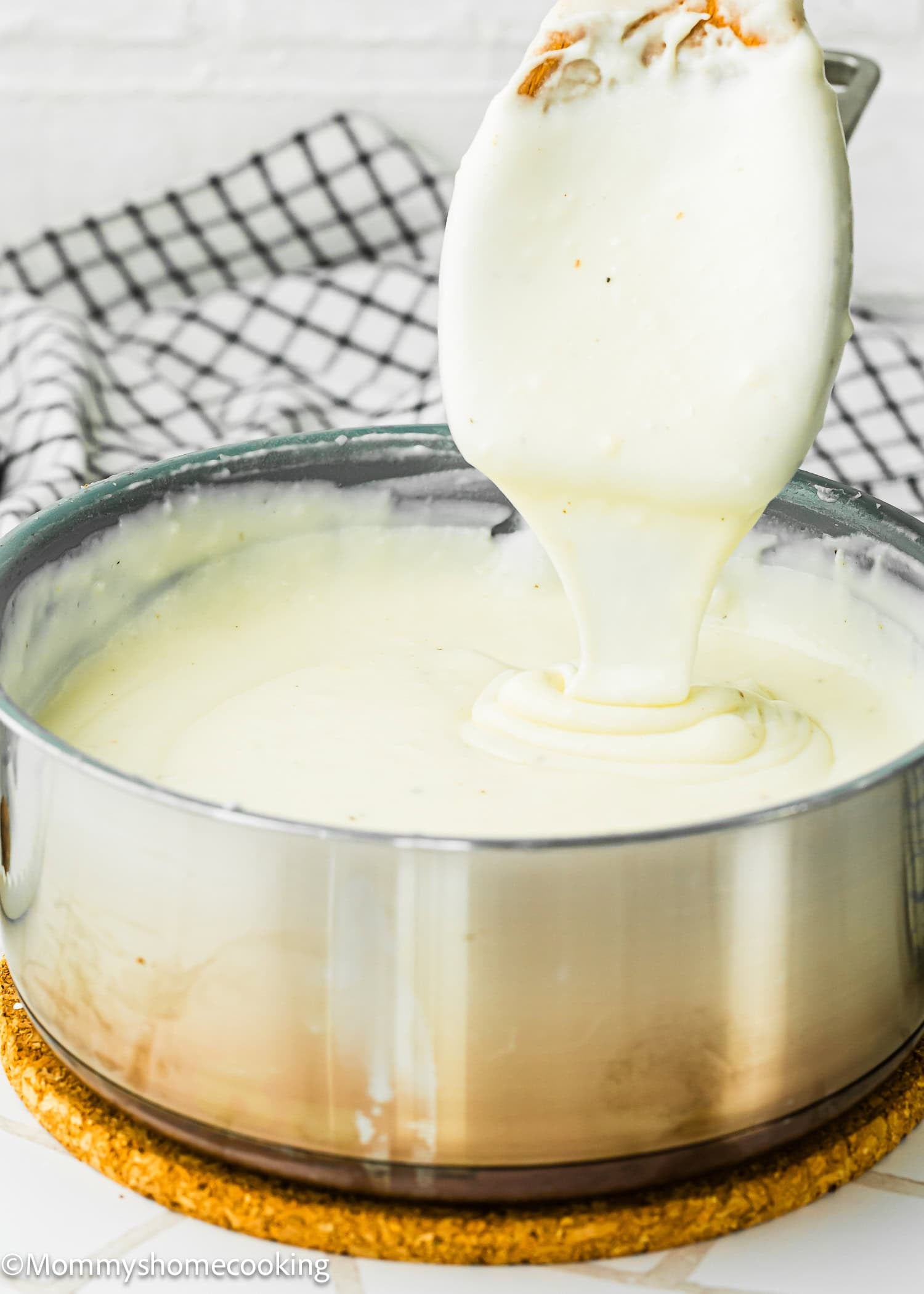 A saucepan filled with Easy Béchamel Sauce (Classic White Sauce Recipe), with a spatula lifting some sauce out, placed on a cork mat with a checkered cloth in the background.