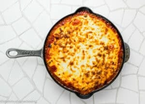 A round cast iron skillet filled with baked, golden-brown Easy Gnocchi Lasagna (with Béchamel sauce) sits on a white tiled surface.