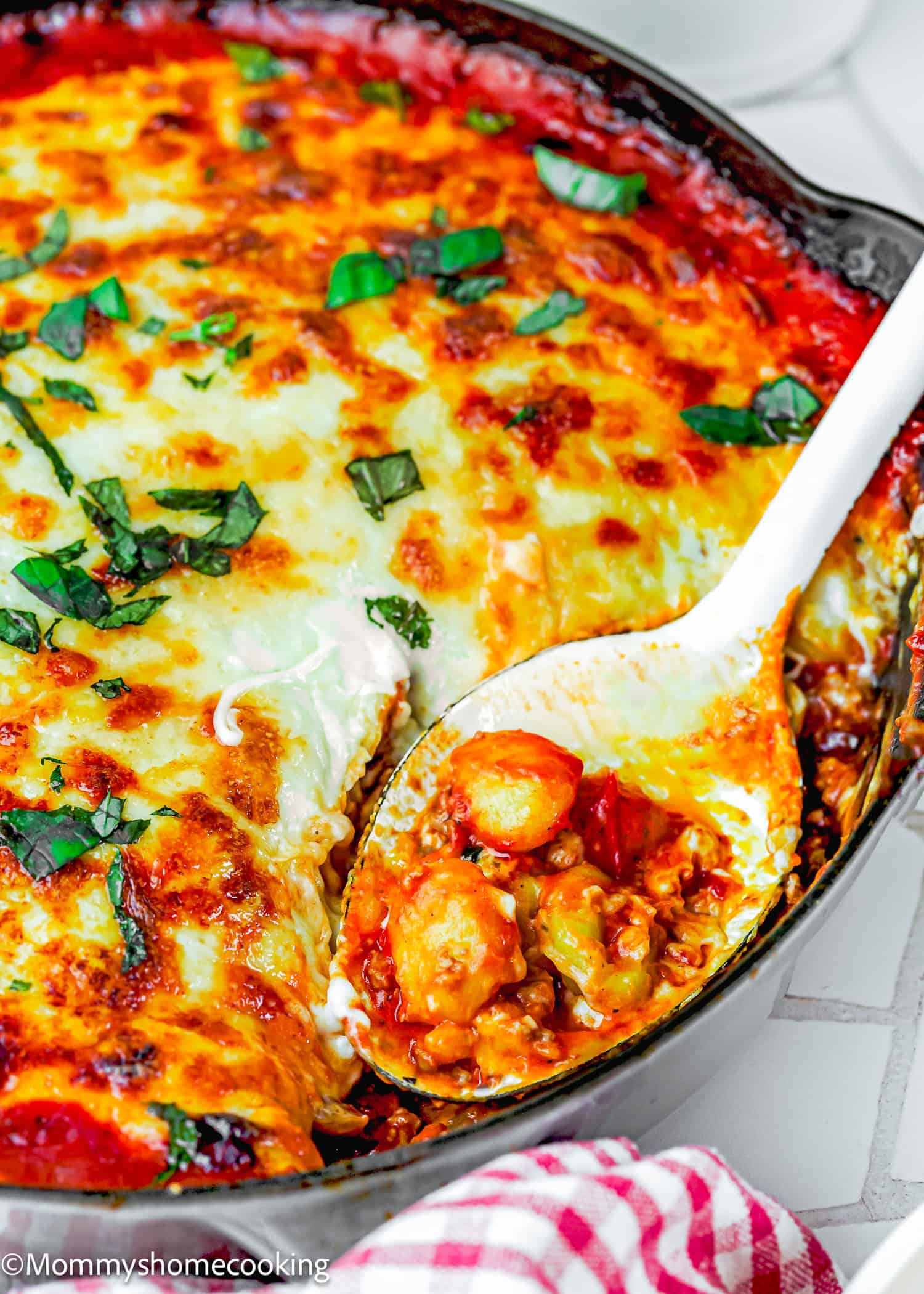 An easy Gnocchi Lasagna with Béchamel sauce, baked in a skillet with melted cheese and tomato sauce, topped with fresh basil. A serving spoon lifts a delicious portion.