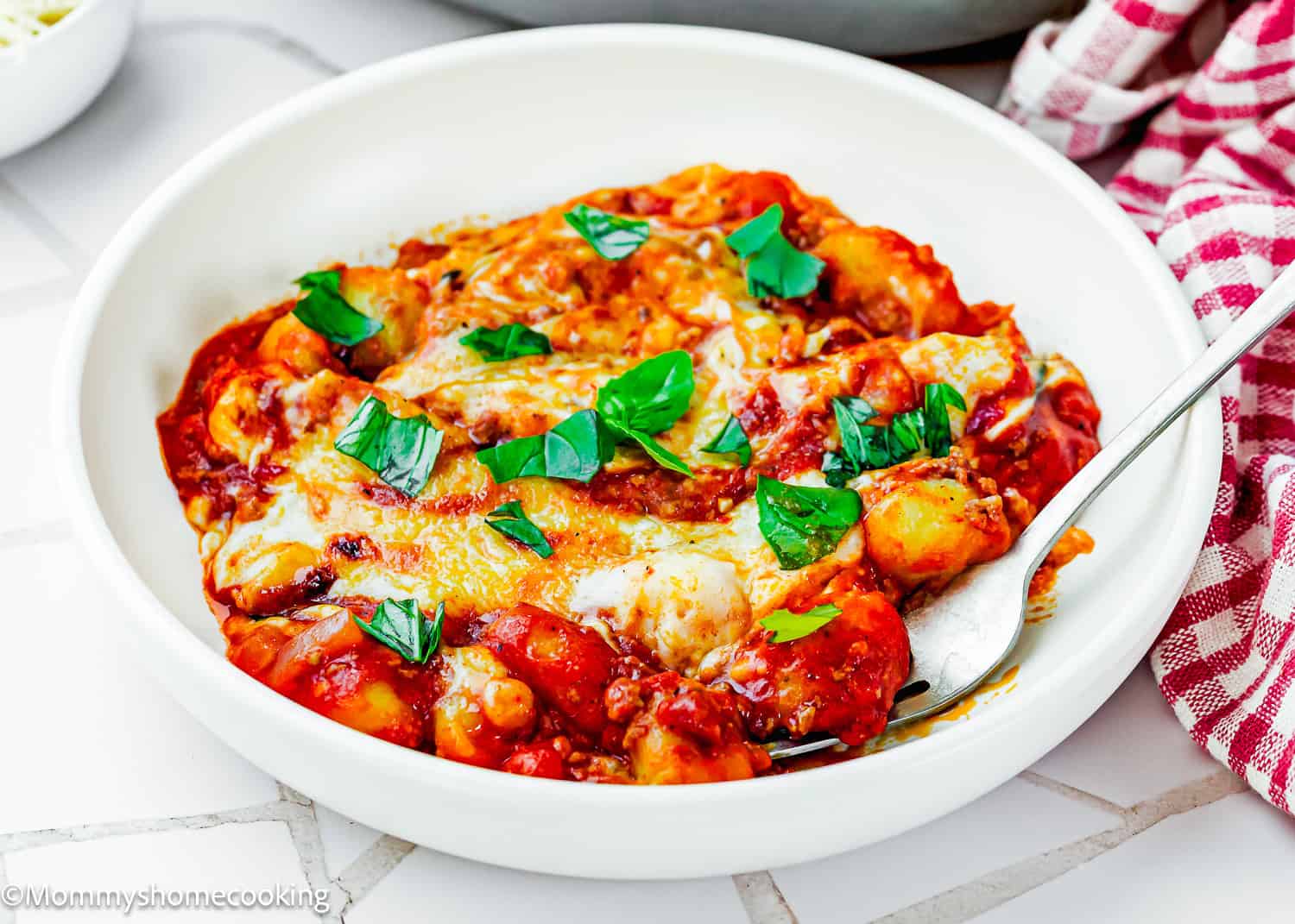 A white bowl with Easy Gnocchi Lasagna in tomato sauce, topped with melted cheese and fresh basil leaves, and a fork on the side.