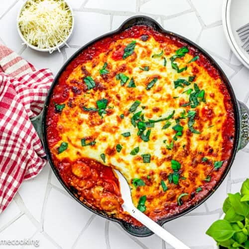 A skillet of easy gnocchi lasagna with béchamel sauce, topped with chopped herbs, sits on a tiled surface, surrounded by a serving spoon, grated cheese, plates, and a red checkered towel.