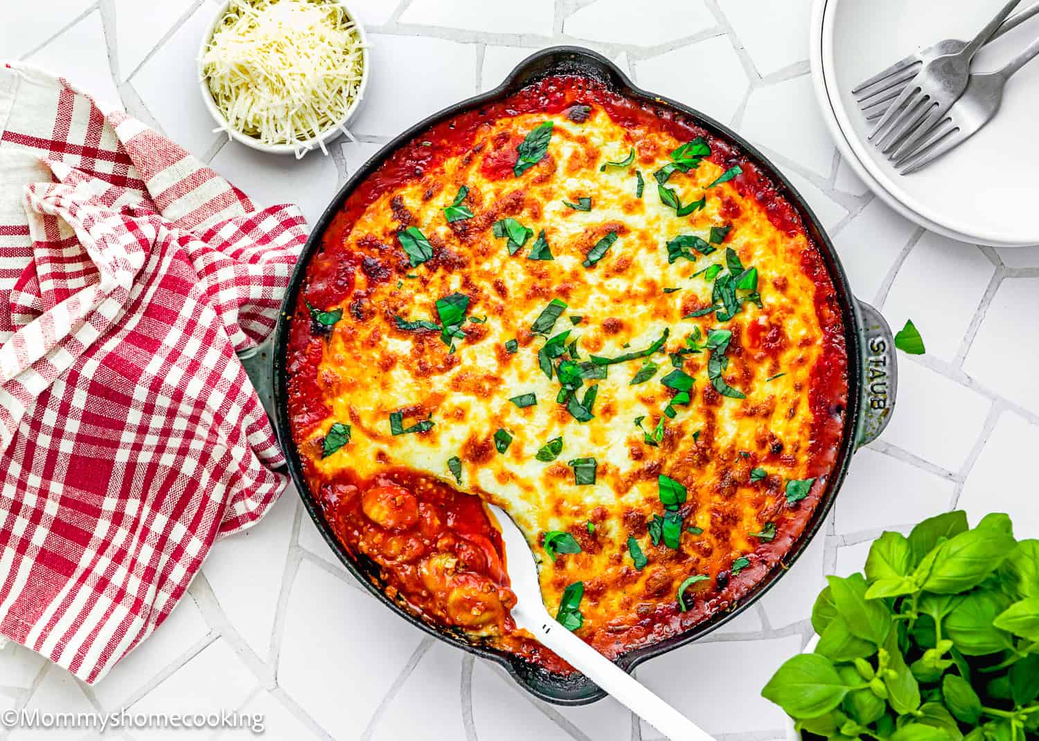 A skillet of easy gnocchi lasagna with béchamel sauce, topped with chopped herbs, sits on a tiled surface, surrounded by a serving spoon, grated cheese, plates, and a red checkered towel.