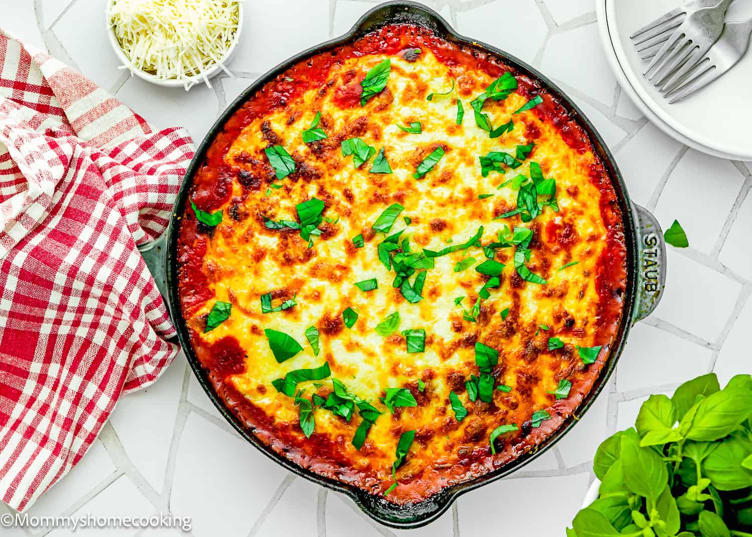 An easy Gnocchi Lasagna (with Béchamel sauce) is baked in a skillet, topped with melted cheese and fresh basil, and sits on a white tiled surface beside a red checkered towel, grated cheese, and plates with forks.
