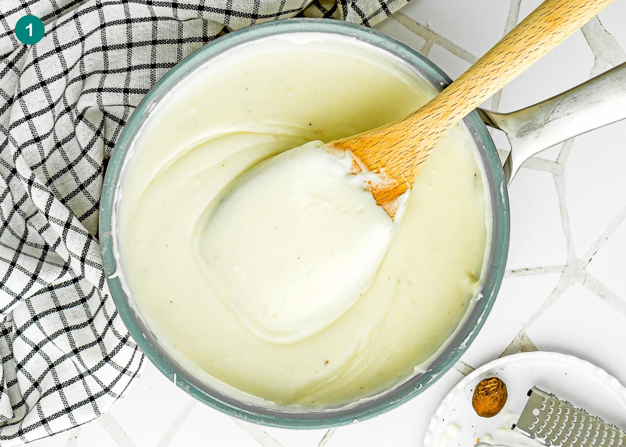 A saucepan filled with smooth, white Béchamel sauce for Easy Gnocchi Lasagna is being stirred with a wooden spoon, next to a checkered kitchen towel on a white tiled surface.