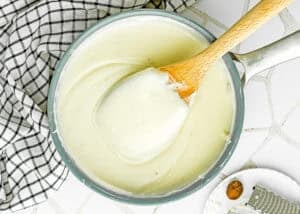 A saucepan filled with creamy béchamel sauce, stirred with a wooden spoon, sits beside a checkered cloth on a tiled surface—perfect for making Easy Gnocchi Lasagna.