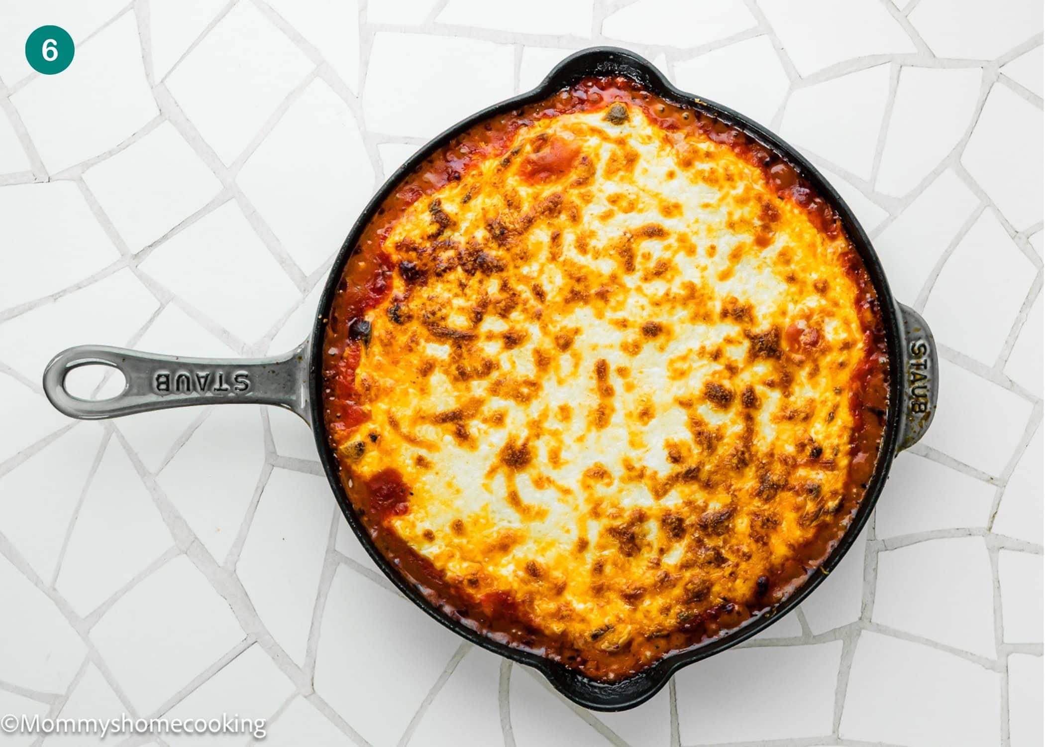 A round cast iron skillet filled with baked, golden-brown Easy Gnocchi Lasagna (with Béchamel sauce) sits on a white tiled surface.