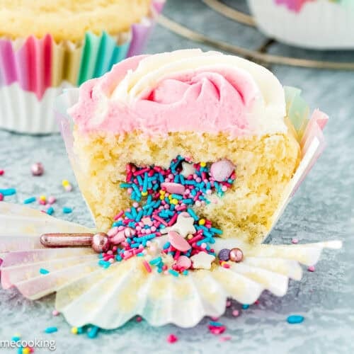 An eggless birthday cupcake with pink and white frosting is cut open, revealing a surprise inside—colorful sprinkles and candy. The cupcake sits on a marble surface with scattered sprinkles around.