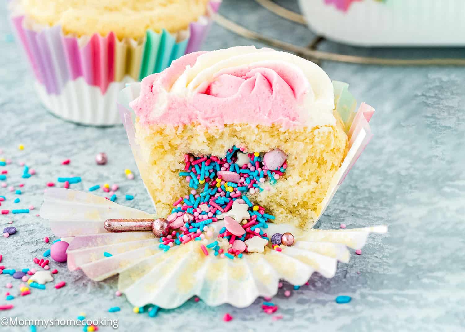 An eggless birthday cupcake with pink and white frosting is cut open, revealing a surprise inside—colorful sprinkles and candy. The cupcake sits on a marble surface with scattered sprinkles around.