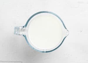 A glass measuring cup filled with milk sits on a white textured surface, perfect for making Eggless Birthday Cupcakes (With Surprise inside).