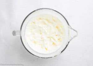 A glass mixing bowl filled with fluffy white whipped cream sits on a white surface, ready to top Eggless Birthday Cupcakes (With Surprise inside).