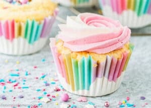 An eggless birthday cupcake in a rainbow-striped wrapper, topped with pink and white swirled frosting and colorful sprinkles—plus a sprinkle surprise inside!.