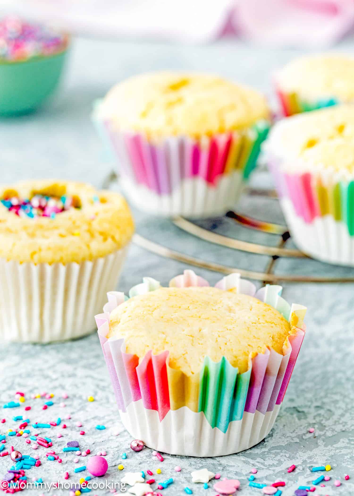 Four Eggless Birthday Cupcakes (With Surprise inside) in colorful paper liners are displayed on a cooling rack and surface, with sprinkles scattered around.