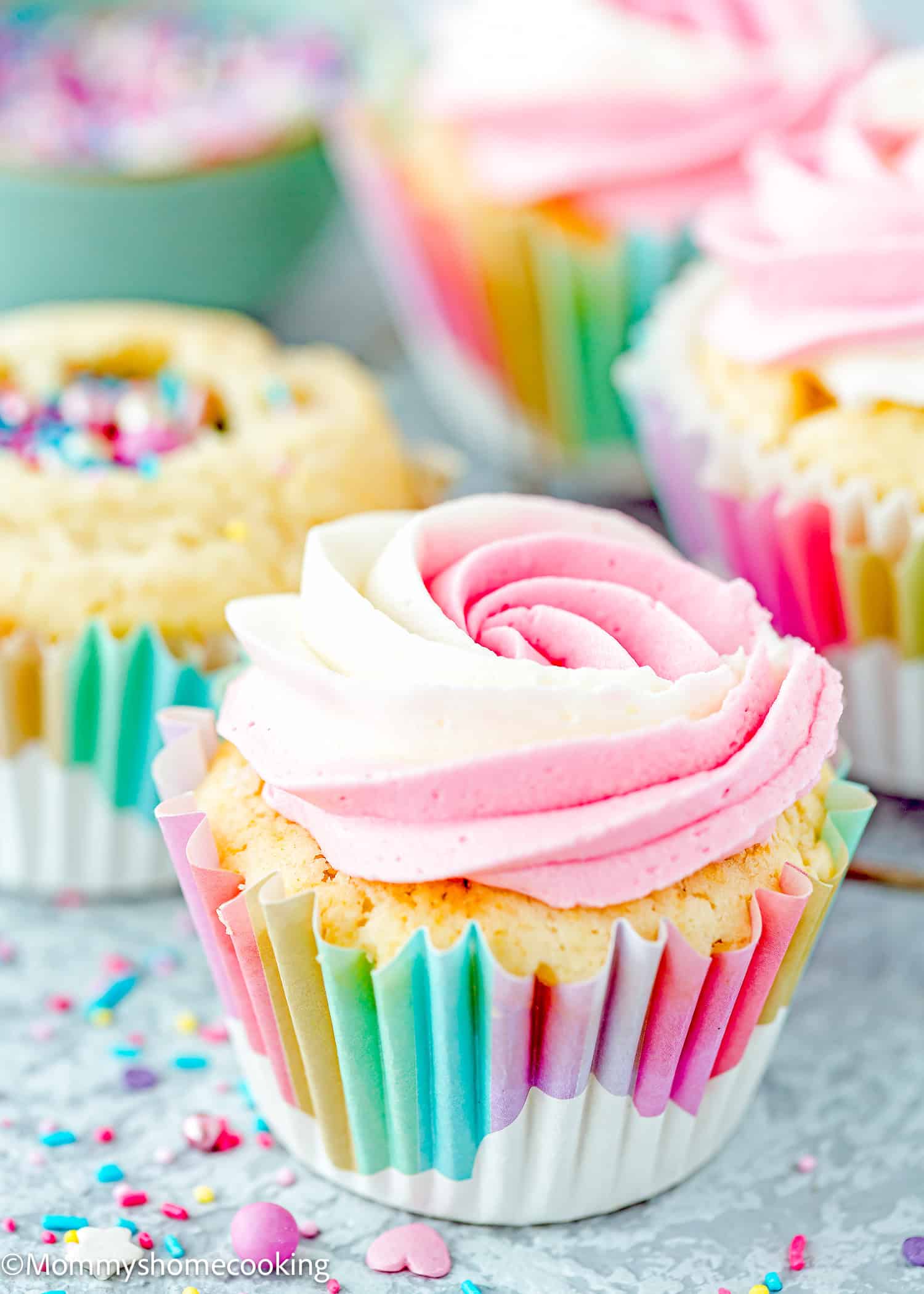 An Eggless Birthday Cupcake (With Surprise inside) sits in a rainbow-striped wrapper, topped with pink and white swirled frosting and surrounded by colorful sprinkles and other decorated cupcakes.