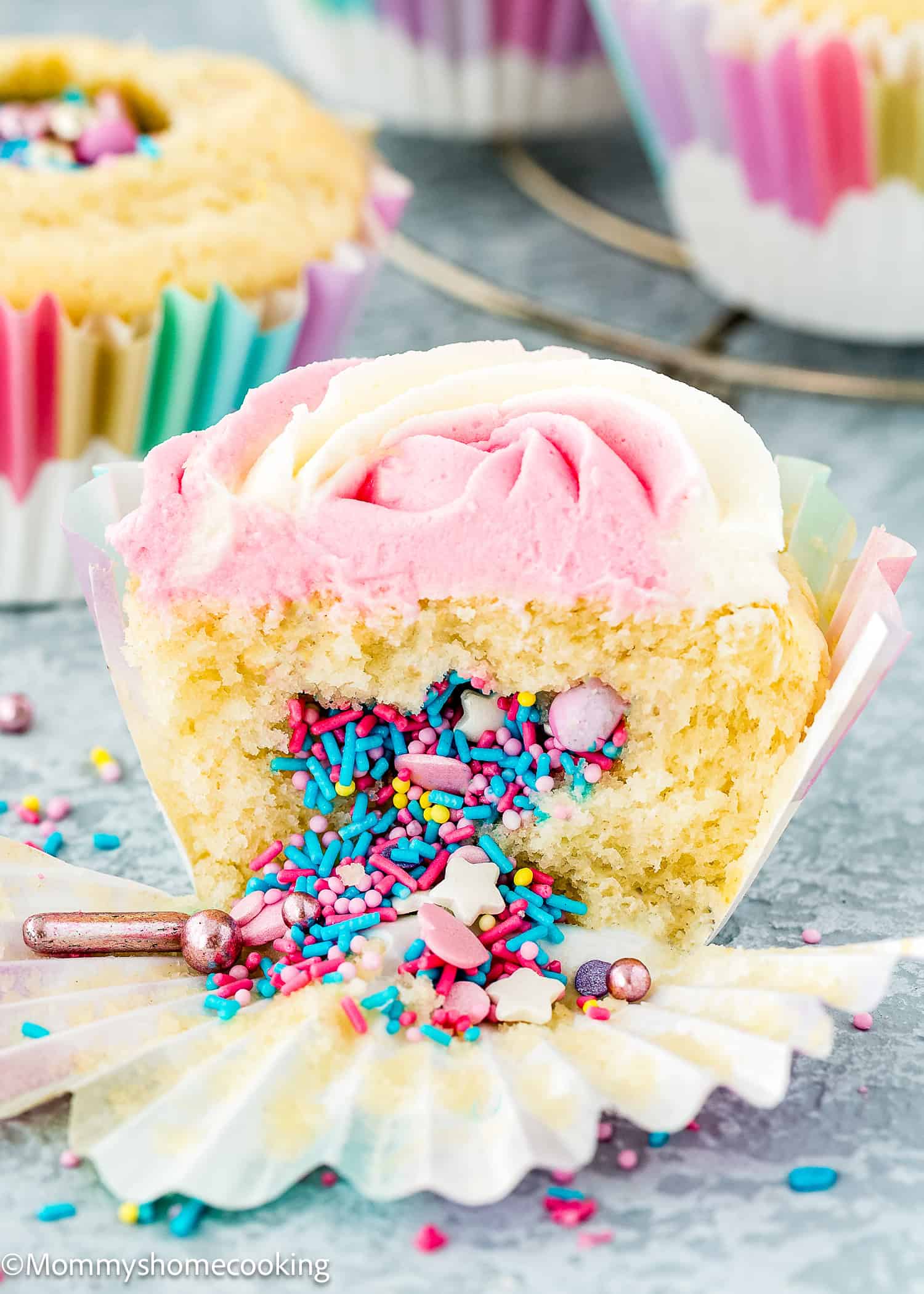 An Eggless Birthday Cupcake (With Surprise inside) is cut open to reveal colorful sprinkles hidden inside its pink and white frosting, sitting in a partially unwrapped paper liner.