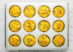 A muffin tin with twelve Eggless Cheesy Savory Muffins in paper liners sits on a white tiled surface.